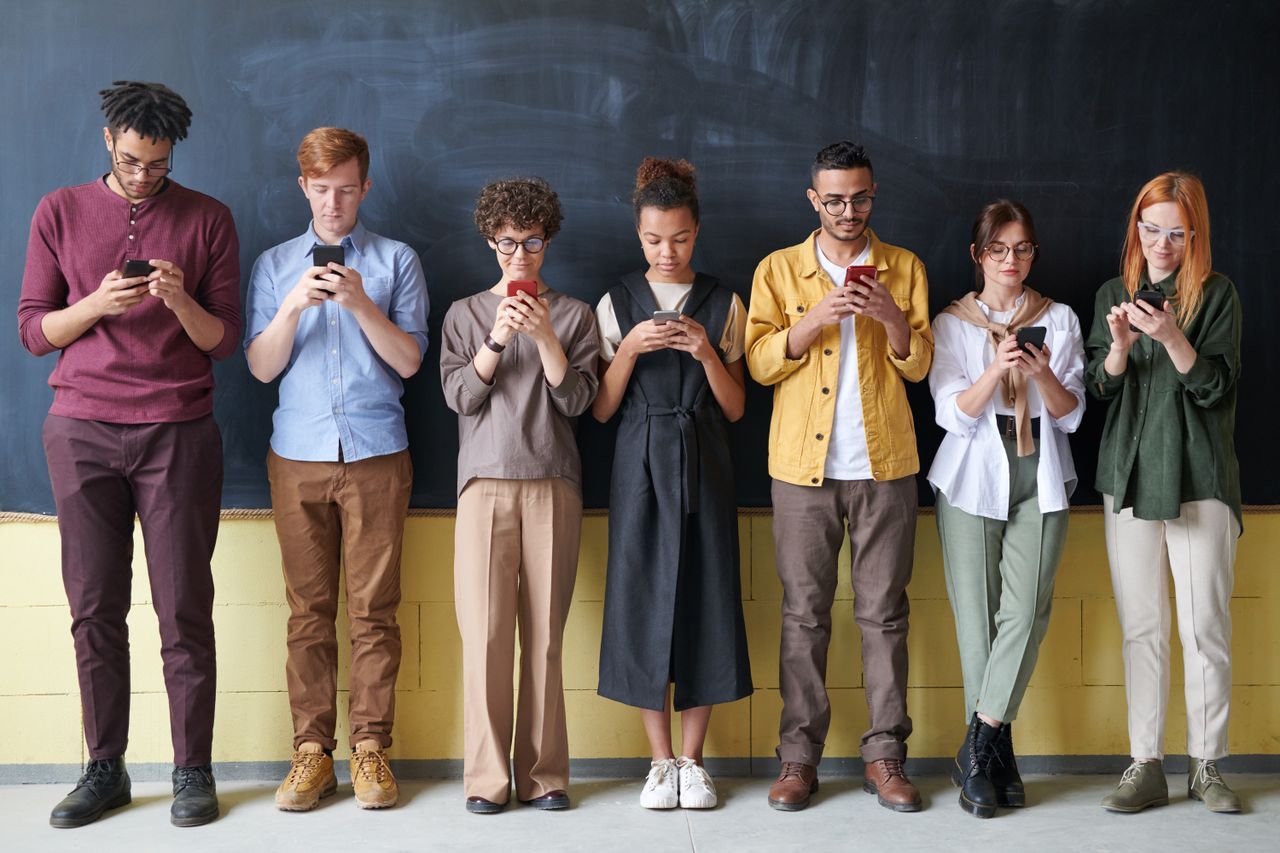Home A diverse group of adults standing in a row, engaged with smartphones, in front of a blackboard.